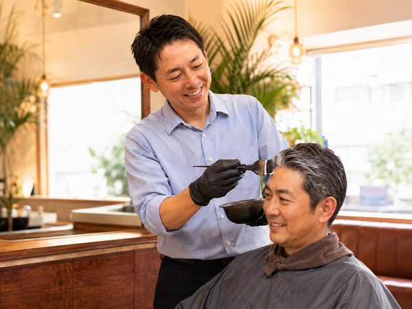 明るく落ち着いた理容室で、笑顔の店主が男性客の白髪にカラー剤を丁寧に塗っている様子。
